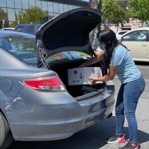 Masked woman loading box of diapers into a car trunk.