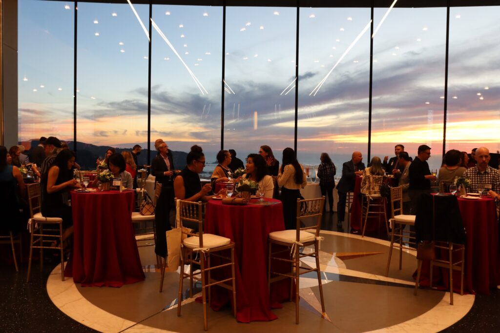 Large dining room with high top tables and people grabbing food in front of a sunset background. 