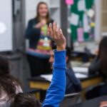 Student in blue sweatshirt raises hand in front of teacher to ask a question.