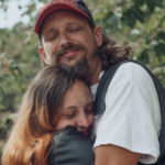 Father in white t-shirt and brown vest hugging his teenage daughter who is wearing a black leather jacket.